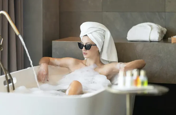 A person with their hair wrapped in a towel and wearing sunglasses is relaxing in a bubble bath inside a bathroom with modern decor. A faucet with running water, toiletries, and folded towels are visible around the bathtub