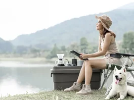 A young woman sitting on a camping chair by a lake, holding a book, with a white dog beside her, a portable coffee maker set on a cooler, and mountains in the background.