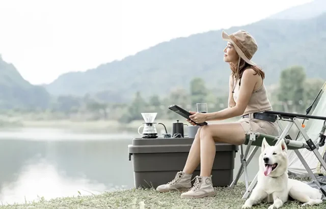 A young woman sitting on a camping chair by a lake, holding a book, with a white dog beside her, a portable coffee maker set on a cooler, and mountains in the background.