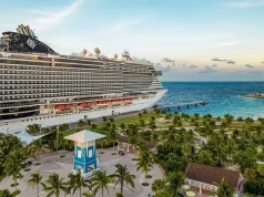 MSC Cruises Offers The Ultimate Winter Escape To The Caribbean’s Most Idyllic Destinations shows a cruise ship docked at a tropical port. The scene features a large, multi-deck cruise ship beside a palm-lined promenade with a lighthouse in the background and the ocean stretching out beyond the port. The setting suggests a popular Caribbean destination, with the ship likely belonging to MSC Cruises based on the logo visible on the vessel. The overall atmosphere is relaxed and idyllic, capturing the essence of a tropical getaway