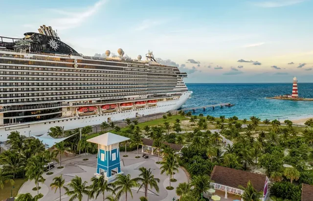 shows a cruise ship docked at a tropical port. The scene features a large, multi-deck cruise ship beside a palm-lined promenade with a lighthouse in the background and the ocean stretching out beyond the port. The setting suggests a popular Caribbean destination, with the ship likely belonging to MSC Cruises based on the logo visible on the vessel. The overall atmosphere is relaxed and idyllic, capturing the essence of a tropical getaway