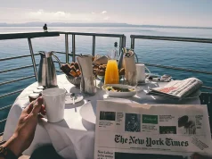 Cruise vs. Land Vacations: Why Cruising Could Be the Perfect Choice for Your 2025 Escape A breakfast table on a deck overlooking calm water, set with coffee, orange juice, pastries, and a newspaper. A person holds a mug while a bird perches on the railing, with distant mountains in the background