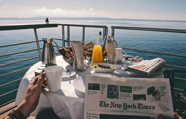 A breakfast table on a deck overlooking calm water, set with coffee, orange juice, pastries, and a newspaper. A person holds a mug while a bird perches on the railing, with distant mountains in the background