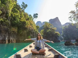 A woman spreading her hands on a boat on a body of water, surrounded by hills and trees.