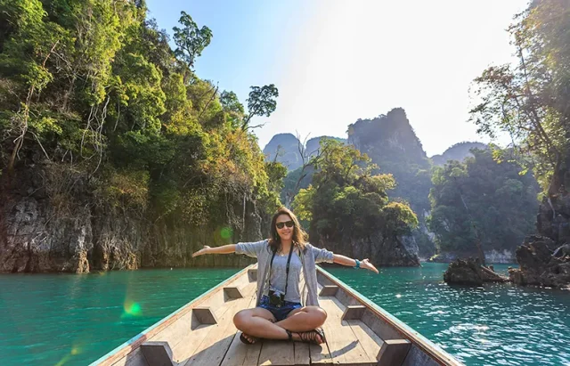 A woman spreading her hands on a boat on a body of water, surrounded by hills and trees.