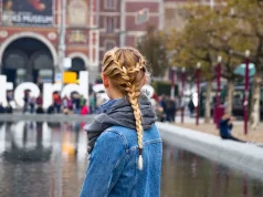 Explore, Play And Learn: The Top Family Friendly European City Breaks Woman with braided hair in a denim jacket stands by water in Amsterdam, with the Rijksmuseum and crowd in the background.