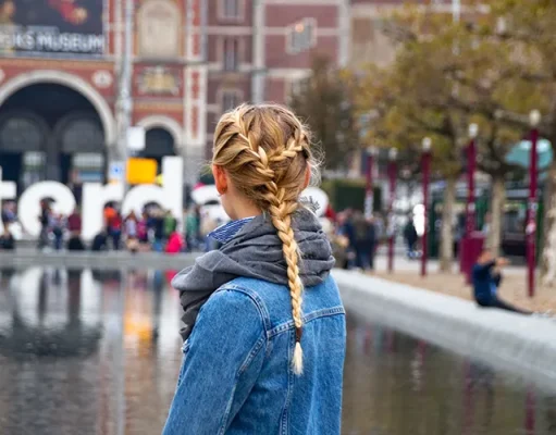 Explore, Play And Learn: The Top Family Friendly European City Breaks Woman with braided hair in a denim jacket stands by water in Amsterdam, with the Rijksmuseum and crowd in the background.