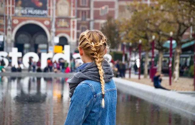 Woman with braided hair in a denim jacket stands by water in Amsterdam, with the Rijksmuseum and crowd in the background.