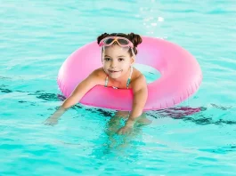 A young girl smiles during her swim lesson at Nemo Swim School, wearing a floatation ring and children’s swim goggles in the pool.