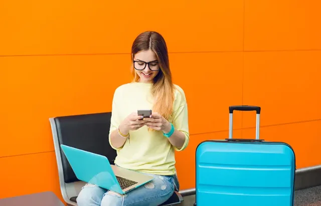 young-girl-with-long-hair-black-glasses-is-sitting-chair-orange-background-she-wears-jellow-sweater-with-jeans-she-has-laptop-knees-suitcase-near-she-is-typing-phone copy