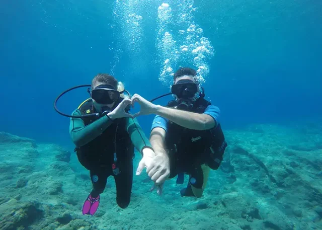 pexels-thanely-104294694-9480734 copy two people scuba diving underwater, making a heart shape with their hands.