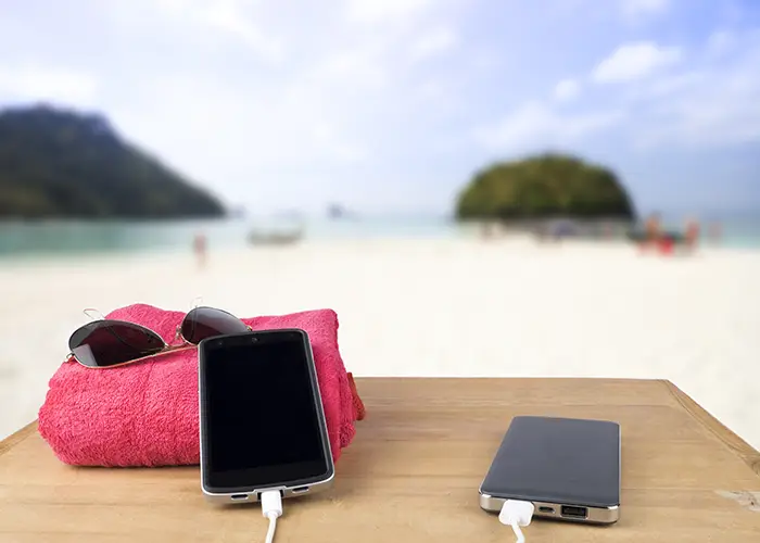 red-tower-sun-glasses-mobile-charging-with-power-bank-wooden-table-blur-beach-sand-blue-sky-background copy