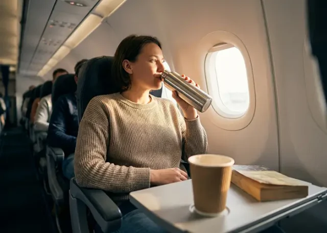 Person in airplane seat enjoying a drink from a thermos.