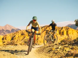 Two mountain bikers cycling on a scenic desert singletrack trail with golden cliffs and mountain views under a clear blue sky