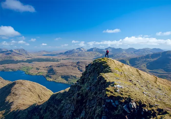 Walker on the South Ridge of Mweelrea Mountain overlooking Killary Fjord in County Mayo, Ireland, with panoramic views of mountains, lakes, and dramatic coastal landscapes.