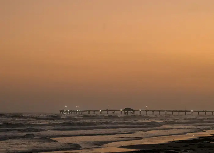 A lit fishing pier stretching into the Gulf at sunset with waves rolling onto the beach