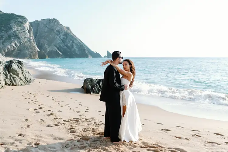 Couple in a white dress and a black suit on Praia da Adraga, with the cliffs and sea stacks of the Sintra coast behind them