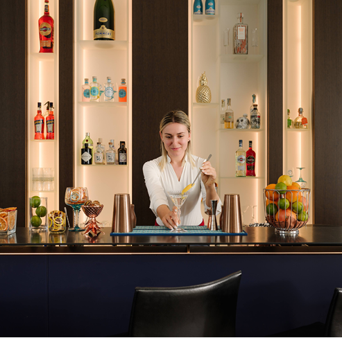 A woman is standing behind a bar with a variety of bottles and fruit. Description generated by AI