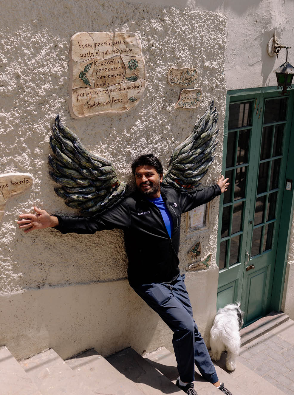 A man with black hair and a blue shirt poses for a picture in front of a wall with angel wings. Description generated by AI