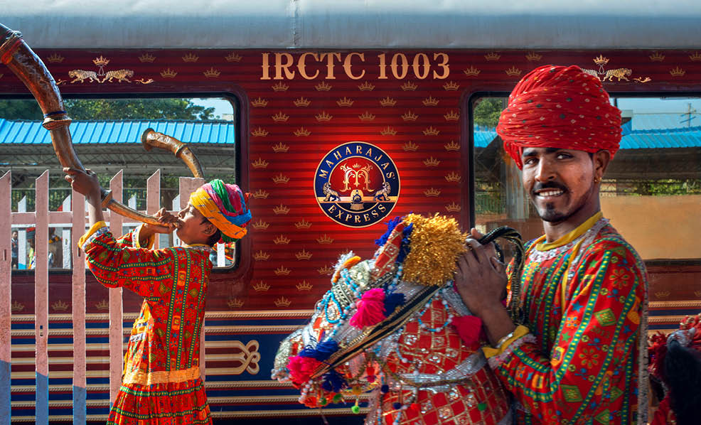 Two men wearing colorful outfits are standing in front of a train. Description generated by AI