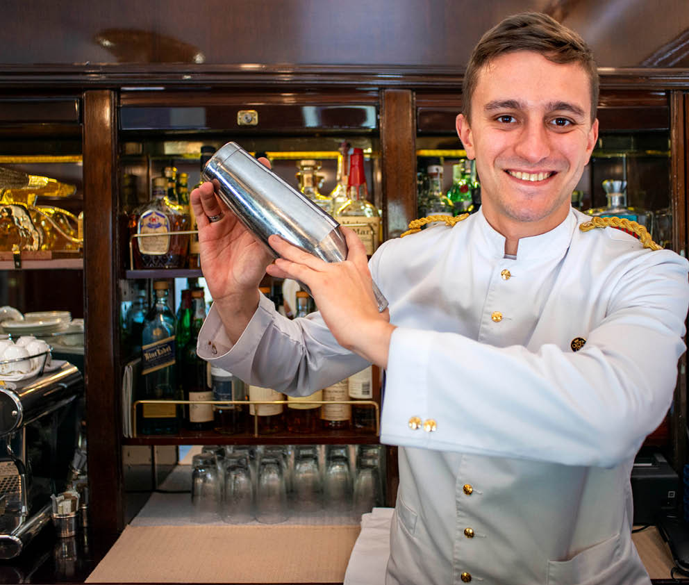 A waiter serves cocktails inside the art deco bar lounge wagon of the train Belmond Venice Simplon Orient Express luxury train.   Sample the finest champagnes in our exclusive bar. Art deco interiors and Lalique glassware conjure the romance and glamour of the Roaring Twenties. Vintages include Taittinger, Laurent Perrier Ros  and Louis Roederer Cristal—perfectly chilled.  An icon of art deco design. Rekindle the romance of the rails as you travel between Europe’s most captivating cities. The Venice Simplon-Orient-Express, or VSOE, is a private luxury train service from London to Venice and other European cities. It is currently owned by Belmond, which operates 45 luxury hotels, restaurants, tourist trains and river cruises in 24 countries. It was agreed in December 2018 for the service to be acquired by LVMH 