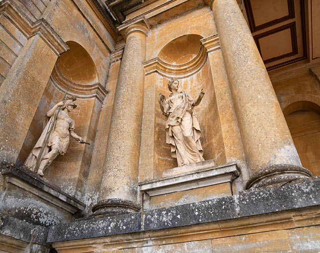 Two classical statues adorn the outer walls of Blenheim Palace, one of a warrior with a spear and the other of a figure in flowing robes, adding to the palace's grandeur.