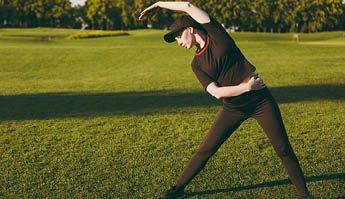 Young athletic girl in black uniform, cap doing sport exercises, warm-up, stretching before running on green lawn in golf course park outdoors on sunny summer day  Fitness, healthy lifestyle concept