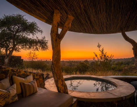 A wooden structure with a hut-like roof and a bench in front of it. The bench has a cushion on it. The sun is setting in the background, casting a warm glow over the scene. AI generated content
