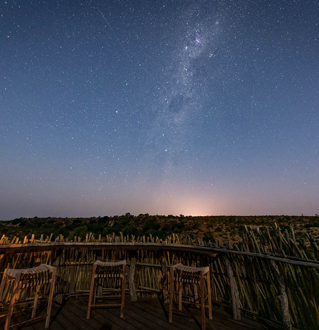 A nighttime scene with a group of chairs arranged on a deck, overlooking a field. The sky is filled with stars, and the Milky Way is visible. AI generated content