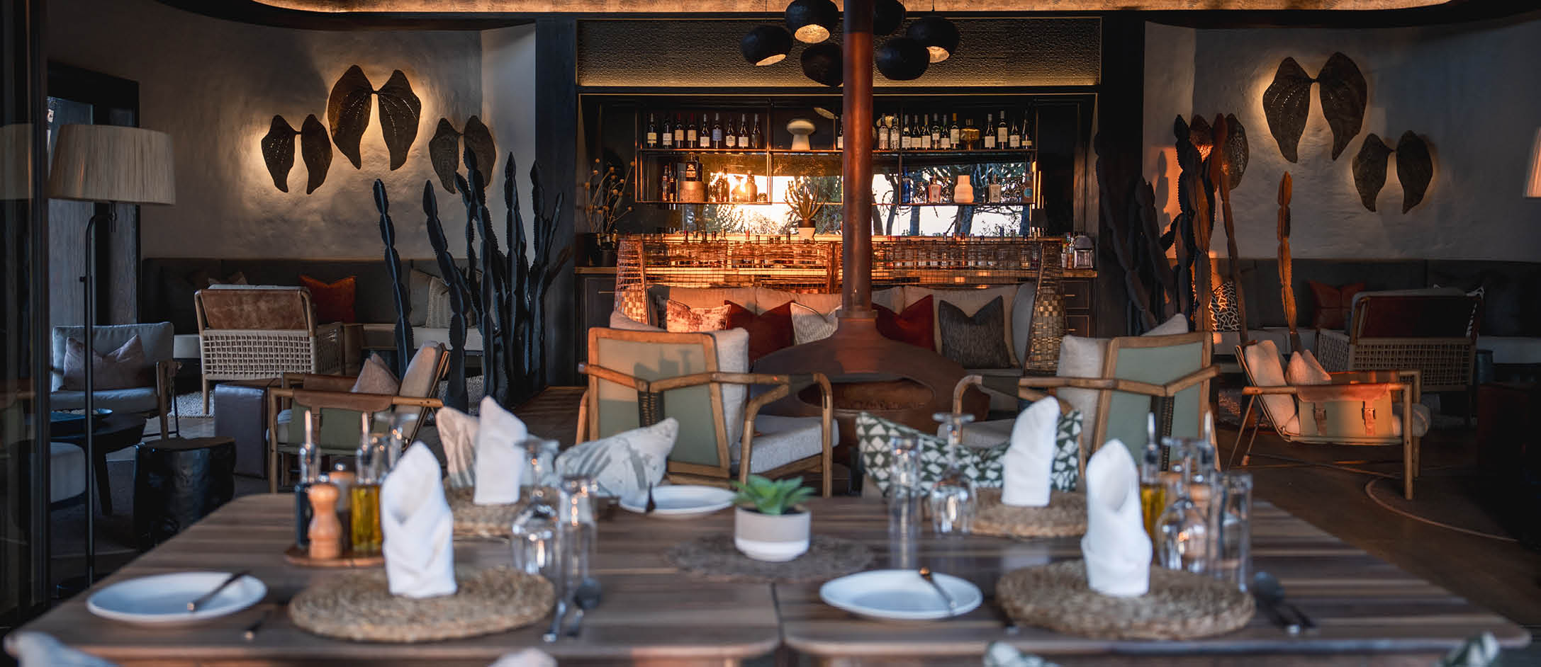 A restaurant with a long dining table set for dinner. The table is set with plates, wine glasses, and utensils. There are several chairs around the table, and a couch in the background. The restaurant has a bar with a bottle on it. AI generated content