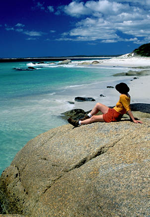 A woman is sitting on a rock near the water, wearing a yellow shirt and a pair of shorts. AI generated content