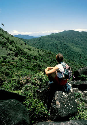 A person is sitting on a rocky hillside, overlooking a valley. They are wearing a backpack and have a handbag nearby. AI generated content