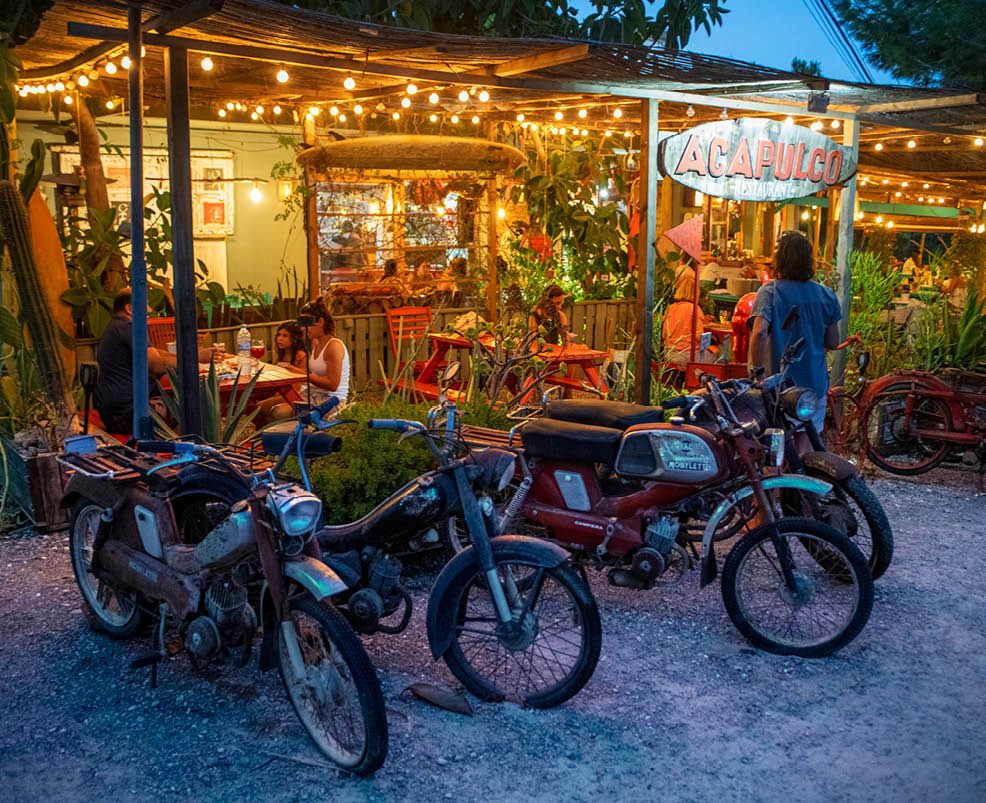 A group of people are sitting at tables outside a restaurant. There are several motorcycles parked nearby, and a few people are standing around them. The scene appears to be a social gathering or a casual meetup. AI generated content