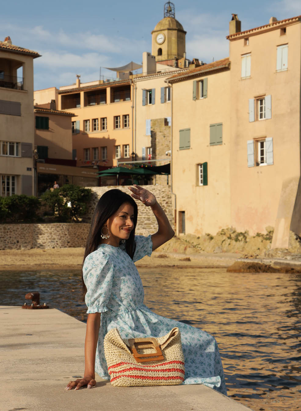 A woman in a flowered dress is standing on a sidewalk near a body of water. She is holding her arm up and appears to be waving. The scene is set in front of a large building. AI generated content