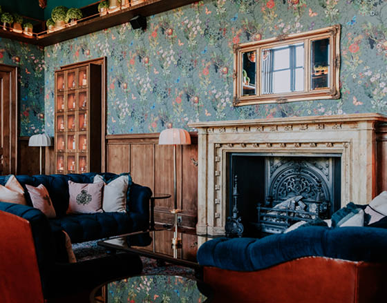 A living room with a couch, chair, and fireplace. The room is decorated with floral wallpaper and has a mirror above the fireplace. AI generated content