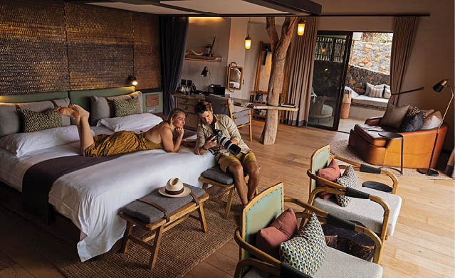 A man and woman are sitting on a bed in a hotel room. The woman is reading a book, while the man is holding a camera. There are two chairs in the room, one near the bed and the other near the window. A suitcase is also present in the room. AI generated content