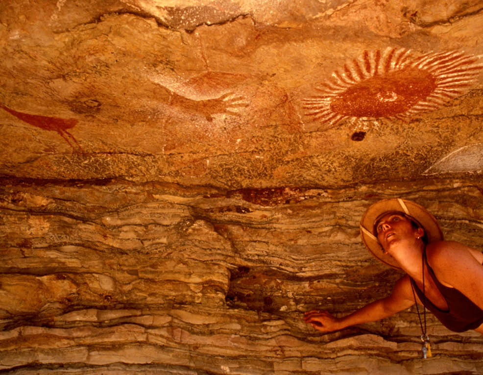 A woman wearing a straw hat is standing in a cave with a painting of a sun on the wall. AI generated content