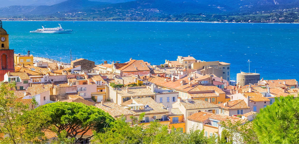 A city overlooks a large body of water, with a boat visible in the distance. The city is surrounded by a lush green hillside, and a lighthouse can be seen in the foreground. AI generated content