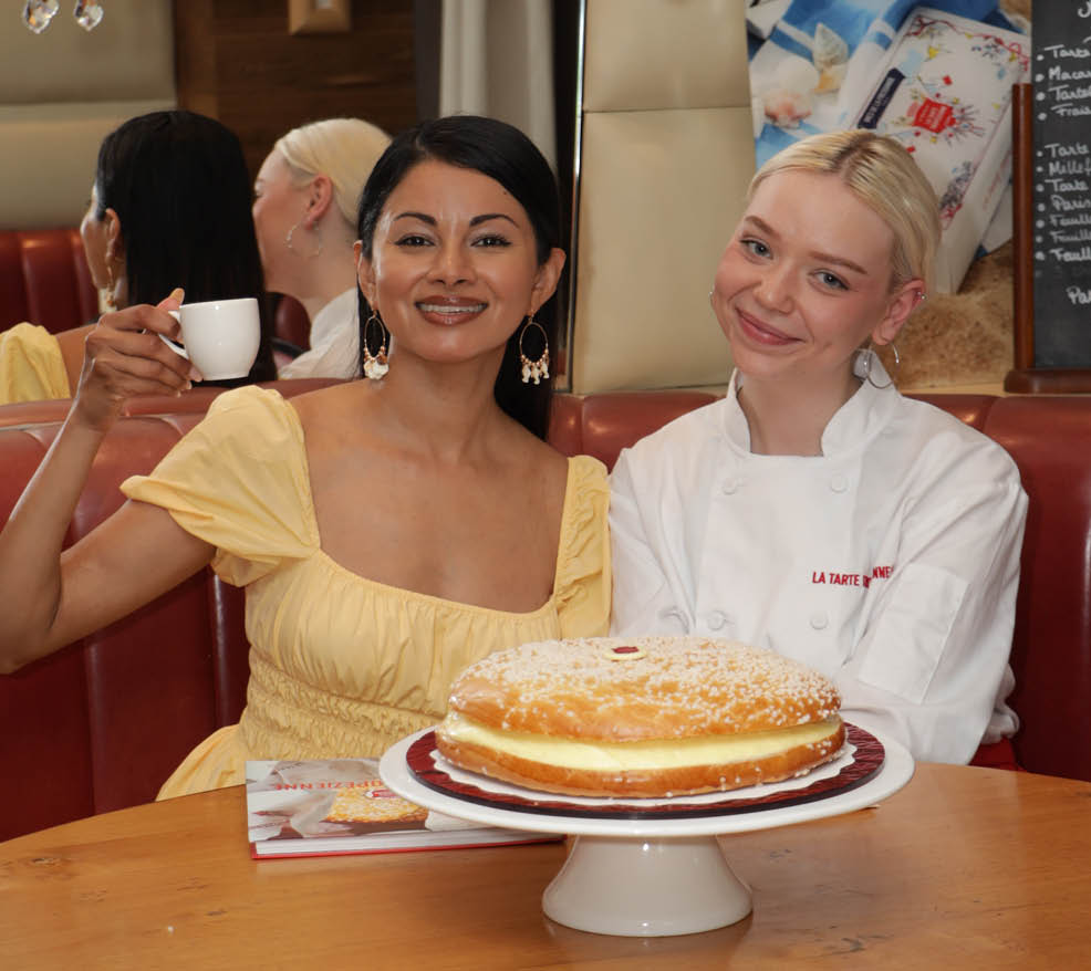 Two women are sitting at a table with a cake in front of them. One woman is wearing a yellow dress and the other is wearing a white shirt. They are both smiling and enjoying their time together. AI generated content