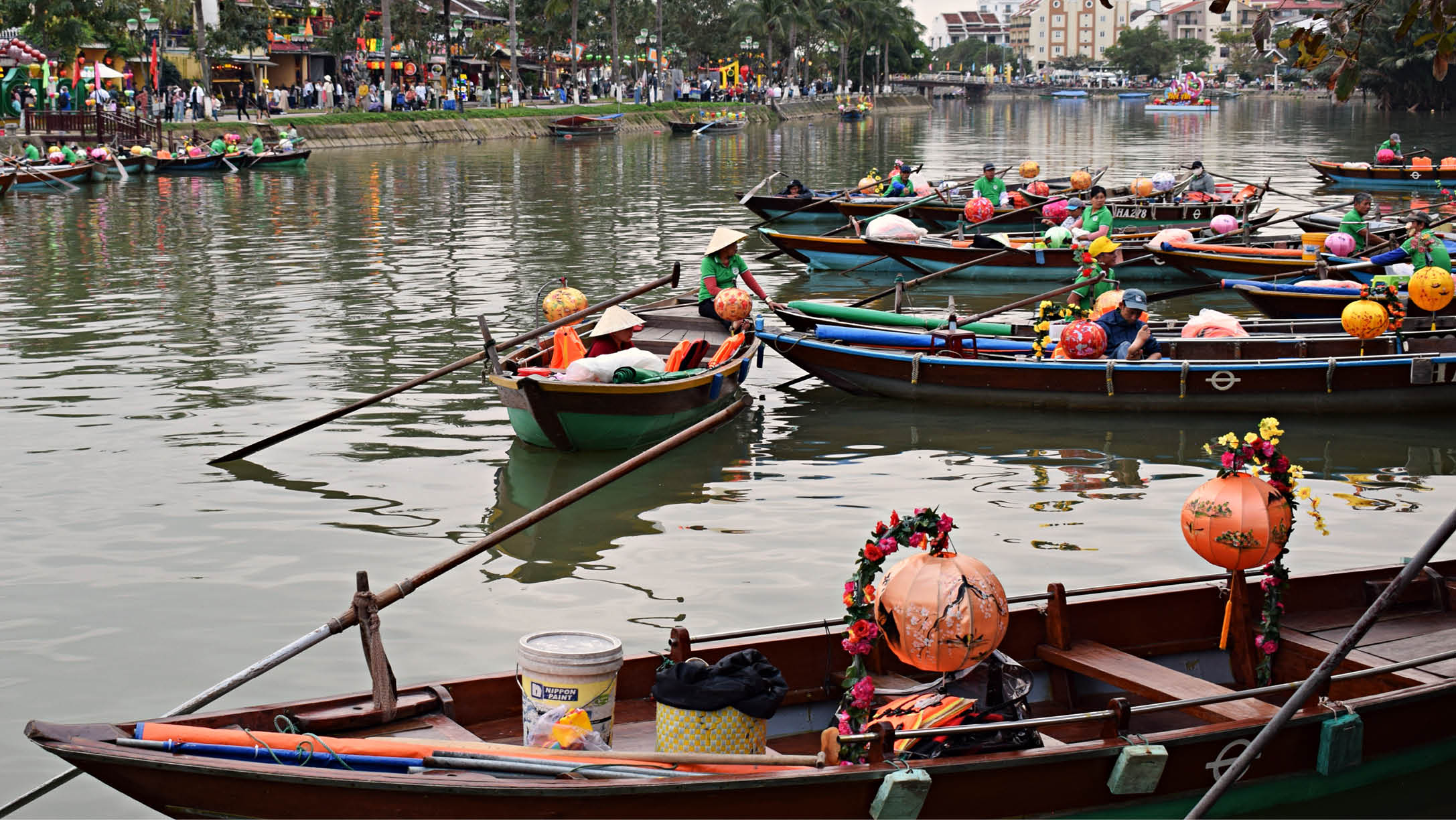 A group of small boats are floating on a river, some with flowers and fruit on them. There are several people in the area, possibly enjoying the view or participating in the event. AI generated content