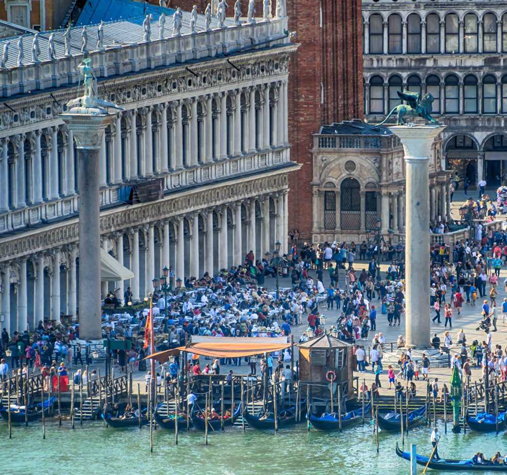 A large crowd of people is gathered in a city square, with many of them walking around and enjoying the day. There are several boats in the water, some of which are docked and others are floating. The scene is bustling with activity, and the people seem to be having a good time. AI generated content