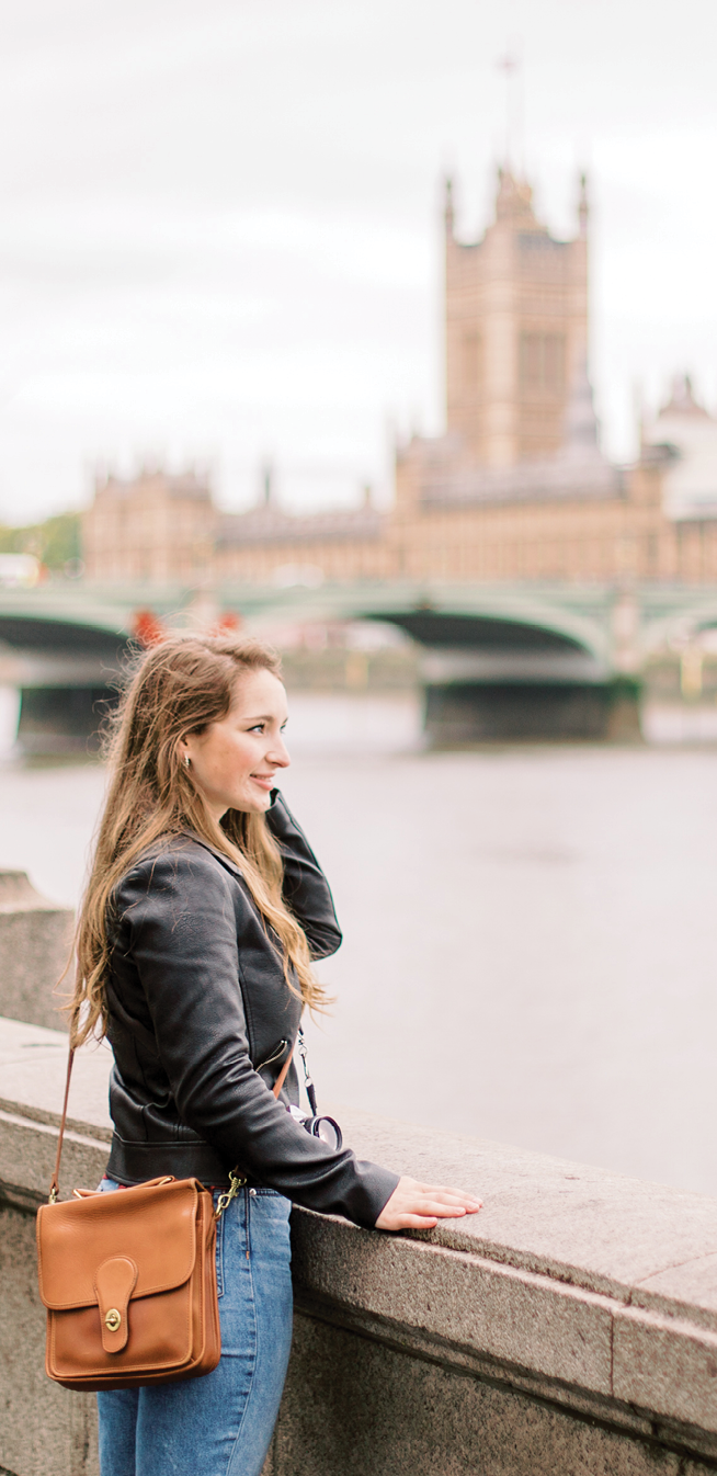 A woman wearing a black jacket is standing on a bridge, looking at the water. She is holding a brown purse. The bridge is overlooking a river, and there are several boats in the water. The scene also includes a clock tower in the background. AI generated content
