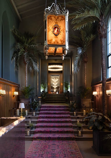 A large, ornate chandelier hangs from the ceiling of a large room. The room features a red carpet and a staircase. There are several potted plants and vases placed throughout the space, adding to the decor. AI generated content