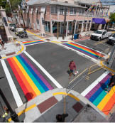 A man walks a dog as Paul Cassidy, right, a Key West Public Works foreman, uses a propane torch to embed thermoplastic strips representing a rainbow flag into asphalt Monday, June 15, 2020, in Key West, Fla. Installation of the rainbow crosswalks capped a project to repave most of Key West’s historic Duval Street. The rainbow flag is a recognized symbol of LGBTQ unity. FOR EDITORIAL USE ONLY (Rob O’Neal/Florida Keys News Bureau/HO)