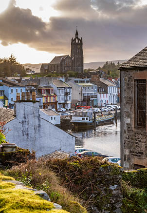 Ancient stone wall covered with moss diagonally in the foreground with a stone house on the left and the Tarbert Free Church cutting the horizon in Argyll and Bute Scotland United Kingdom