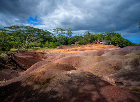 Chamarel seven coloured earth, Black River District, Republic of Mauritius. The seven Coloured Earths are a geological formation and prominent tourist attraction found in the Chamarel plain of the Rivi re Noire District in south-western Mauritius. It is a relatively small area of sand dunes comprising sand of seven distinct colours (approximately red, brown, violet, green, blue, purple and yellow). The main feature of the place is that since these differently coloured sands spontaneously settle in different layers, dunes acquire a surrealistic, striped colouring. Since the earth was first exposed, rains have carved beautiful patterns into the hillside, creating an effect of earthen meringue.